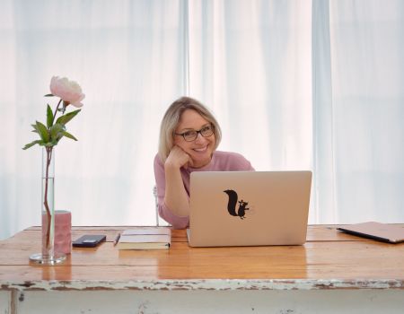 Sabine Läpple sitzt mit Laptop am Schreibtisch und blickt lächelnd in die Kamera – professionelles Branding-Foto für ihre Website.